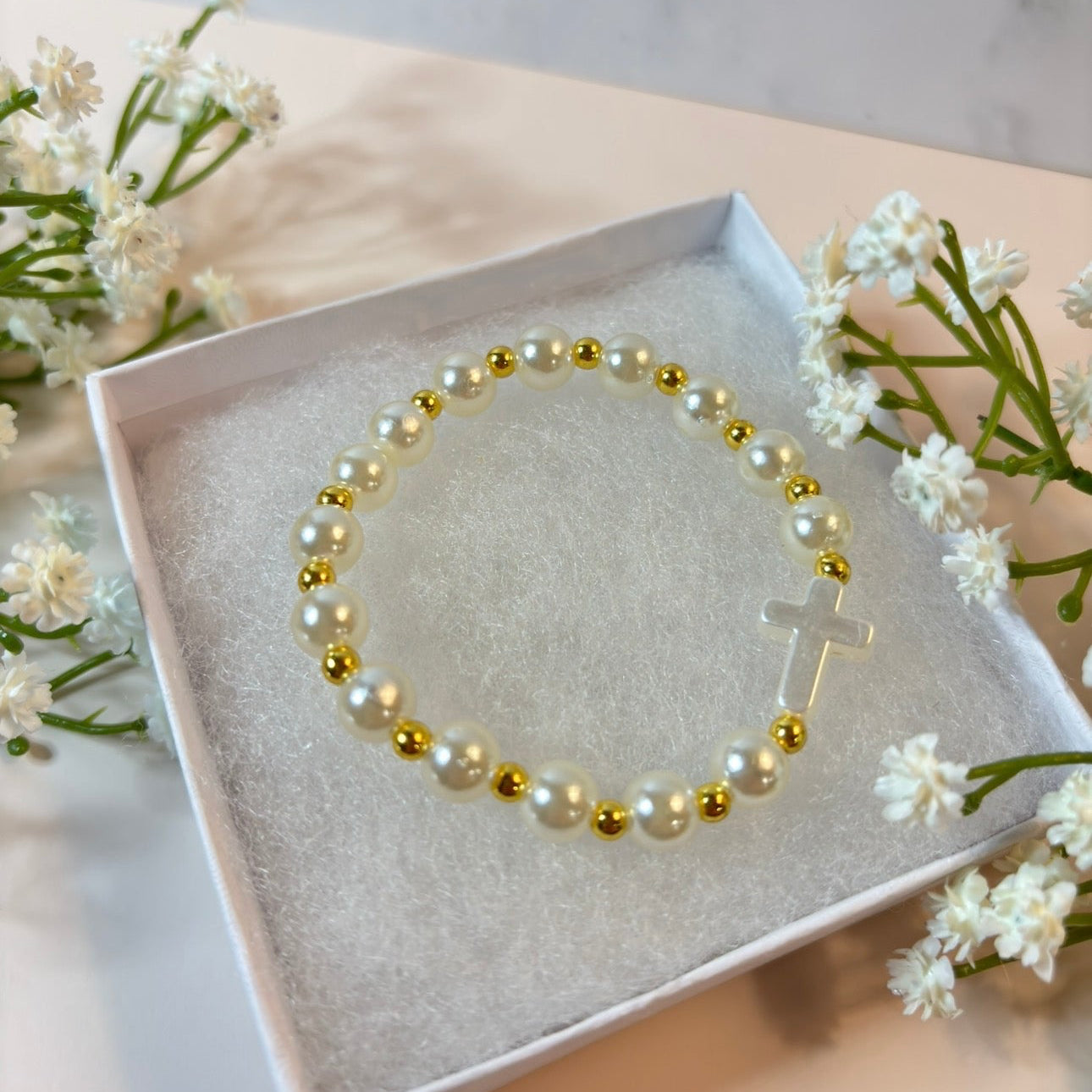 Pearl and gold beaded bracelet with a cross charm in a box, surrounded by white flowers.